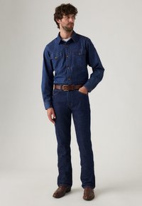 Camisa de mezclilla de manga larga, con dos bolsillos en el pecho, jeans azules, cinturón de cuero marrón y botas marrones. Color azul oscuro y diseño clásico.
