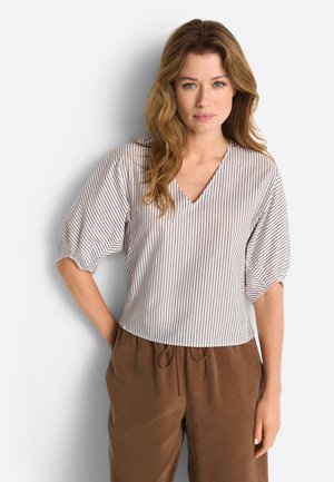 Naised laineliste juustega, kes kannab valget pluusi õhukeste vertikaalsete triipudega ja pruune lahtisi pükse, seisab ühe käega taskus.