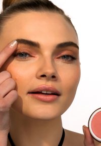 Modell med lys hud og langt hår, som legger makeup med den ene hånden, mens hun holder en rosa blush-beholder med den andre. Enkelt bakgrunn.