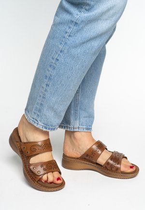 Füße in hellblauen Jeans, die braune Ledersandalen mit dekorativen Ausschnitten tragen, und rote Nagellack auf den Zehen.