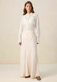 White lace blouse with high neck and long sleeves, paired with a light beige maxi skirt; features ruffled cuffs and a flowing silhouette.