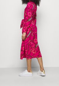 Kvinna klädd i en ljusrosa midiklänning med kedje- och bältemönster, tillsammans med vita sneakers med gyllene klackaccent, stående på sidan.