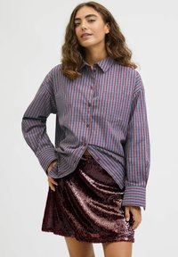 Chemise à rayures rouges et bleues à boutons et manches longues, associée à une mini-jupe en sequins bordeaux brillants, mettant en valeur des textures lisses et un contraste.