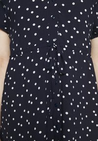 Abito navy con pois bianchi, maniche corte, chiusura con bottoni davanti e una cintura in vita per la regolazione. Tessuto dalla consistenza liscia.