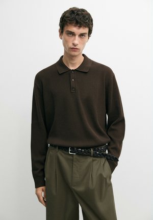 Uomo con capelli corti e ricci che indossa una polo a maniche lunghe marrone scuro, pantaloni plissettati verde oliva e una cintura intrecciata nera.