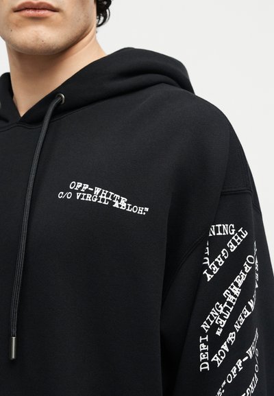 Personne portant un sweat à capuche Off-White noir avec un texte blanc sur la poitrine et un motif de texte circulaire sur la manche droite, sur un fond clair uni.