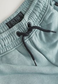 Schmal geschnittene Jogginghose aus hellblauem Stoff mit einem strukturierten Bund, verstellbarem schwarzen Kordelzug und schwarzen Etikettendetails. Weiche Oberfläche.