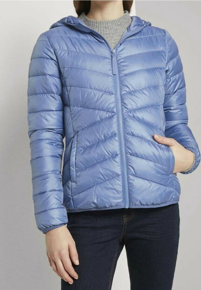 Personne portant une veste matelassée à capuche bleu clair, un pull gris en dessous, un jean foncé, avec une main dans la poche de la veste.