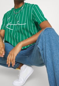 Zielona koszulka z krótkim rękawem w białe paski i haftowanym logo, połączona z jasnoniebieskimi dżinsami i białymi trampkami. Widoczny srebrny łańcuszek.