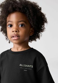 Juodas medvilninis marškinėliai su apvalia iškirpte, su "ALLSAINTS" logotipu žalia spalva ir papildomu tekstu žemiau. Subjekto plaukai garbanoti ir juodi.