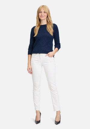 Navy sweater with long sleeves, paired with white skinny jeans and black high heels. Model stands with one hand in pocket.
