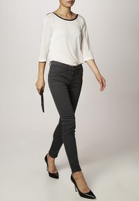 Vit långärmad topp med svart krage, kombinerad med svarta slim-fit jeans och svarta högklackade skor. Modell som håller i en svart clutch.