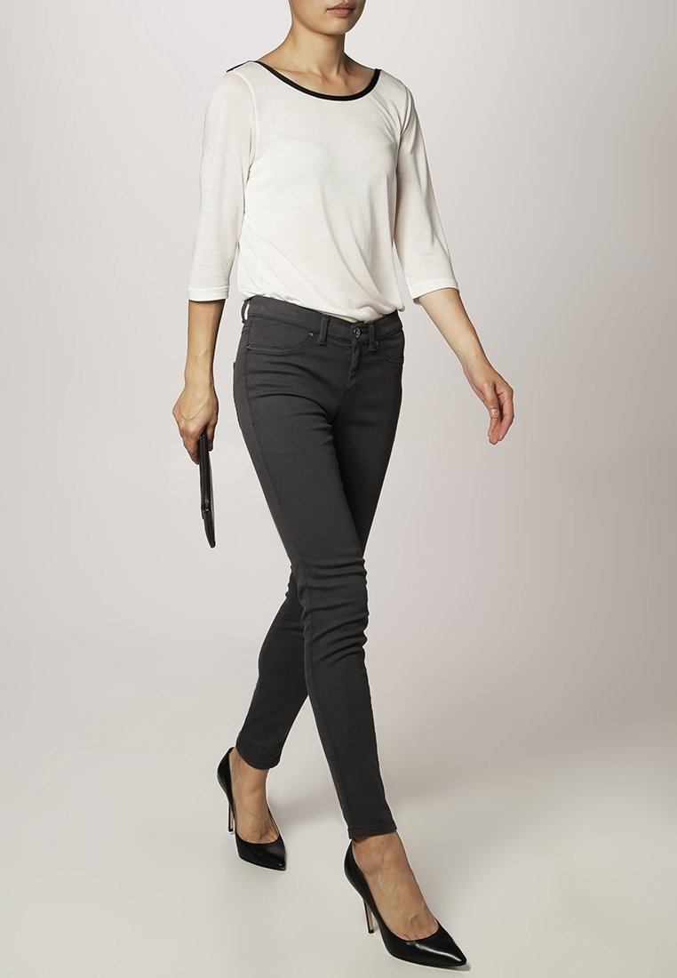 Vit långärmad topp med svart krage, kombinerad med svarta slim-fit jeans och svarta högklackade skor. Modell som håller i en svart clutch.