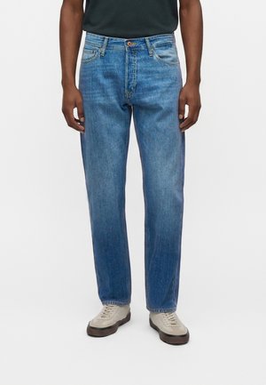 Mann trägt gerade geschnittene blaue Jeans und hellbeige Sneaker, steht vor weißem Hintergrund.