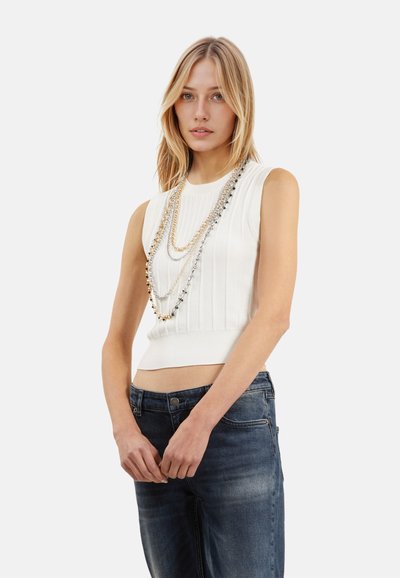 Hvid, ærmeløs striktop med ribbet tekstur og cropped design, parret med denimbukser. Har lagdelte metalliske kæder som tilbehør.