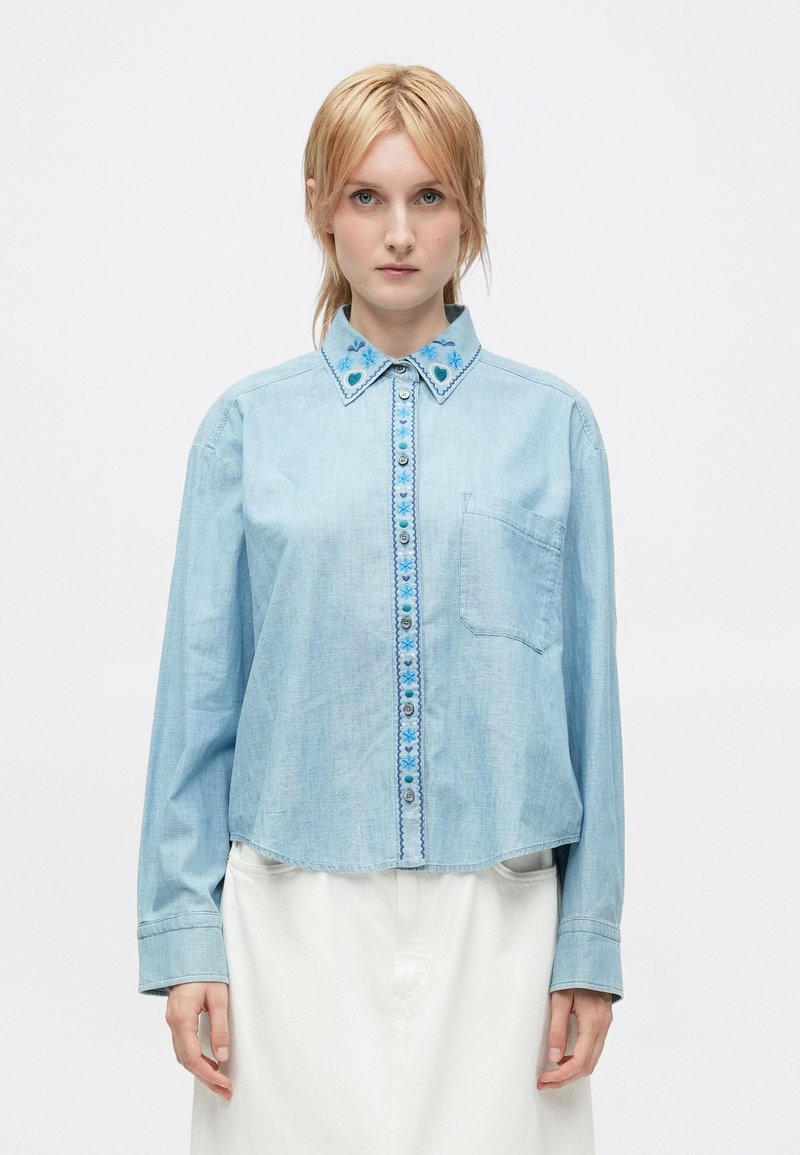 Una donna con capelli biondi indossa una camicia di jeans azzurro chiaro con ricami floreali blu sul collare e sulla linea dei bottoni, abbinata a pantaloni bianchi.