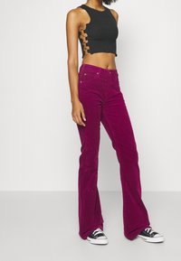 Kvinna i svart crop top med sidokutts, högmidjade lila kordflorsbyxor och svart-vita sneakers som står mot en neutral bakgrund.