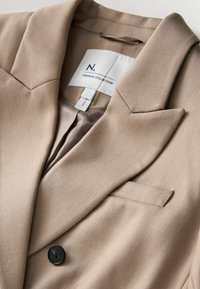 Sacou bej din țesătură texturată, cu guler decupat, nasture negru simplu și detalii discrete cusute. Eticheta interioară este vizibilă.