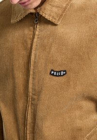 Veste en velours côtelé marron avec fermeture éclair et un patch logo Volcom noir sur le côté gauche. Tissu texturé avec des nervures verticales.