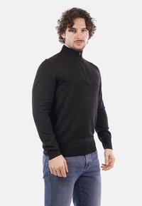 Felpa nera con zip al collo realizzata in un morbido tessuto a maglia. Presenta un design aderente, maniche lunghe e polsini e orlo a coste.