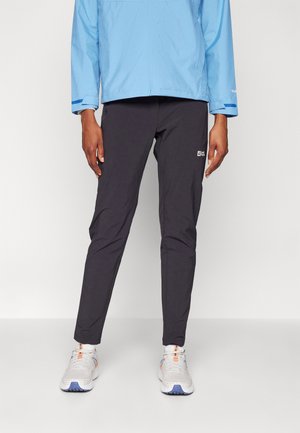 Personne portant un pantalon noir slim, une veste bleu clair et des chaussures de sport blanches, debout devant un fond uni.
