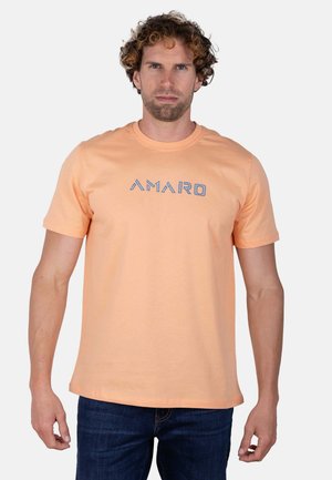 Hombre de pelo rizado con una camiseta color melocotón con "AMARO" en letras azules y vaqueros oscuros, de pie frente a un fondo claro y liso.