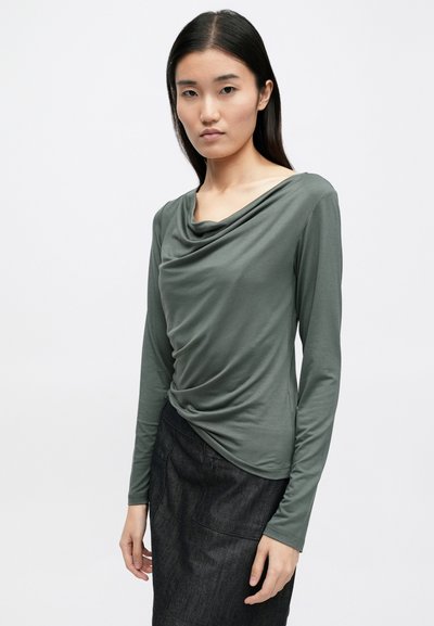 T-shirt de mangas longas em tecido verde suave com decote drapeado e silhueta ajustada, combinado com uma saia escura e texturizada.