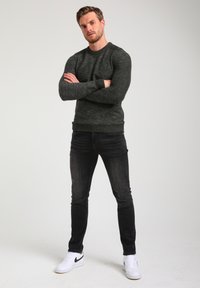 Pull en tricot texturé vert foncé avec un col rond, associé à un jean noir ajusté et des baskets blanches. Tenue décontractée avec des couleurs unies.