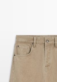 Pantalon en denim beige avec passants pour ceinture, fermeture par bouton métallique, poche avant avec une petite poche pour pièces, et détails de couture visibles.