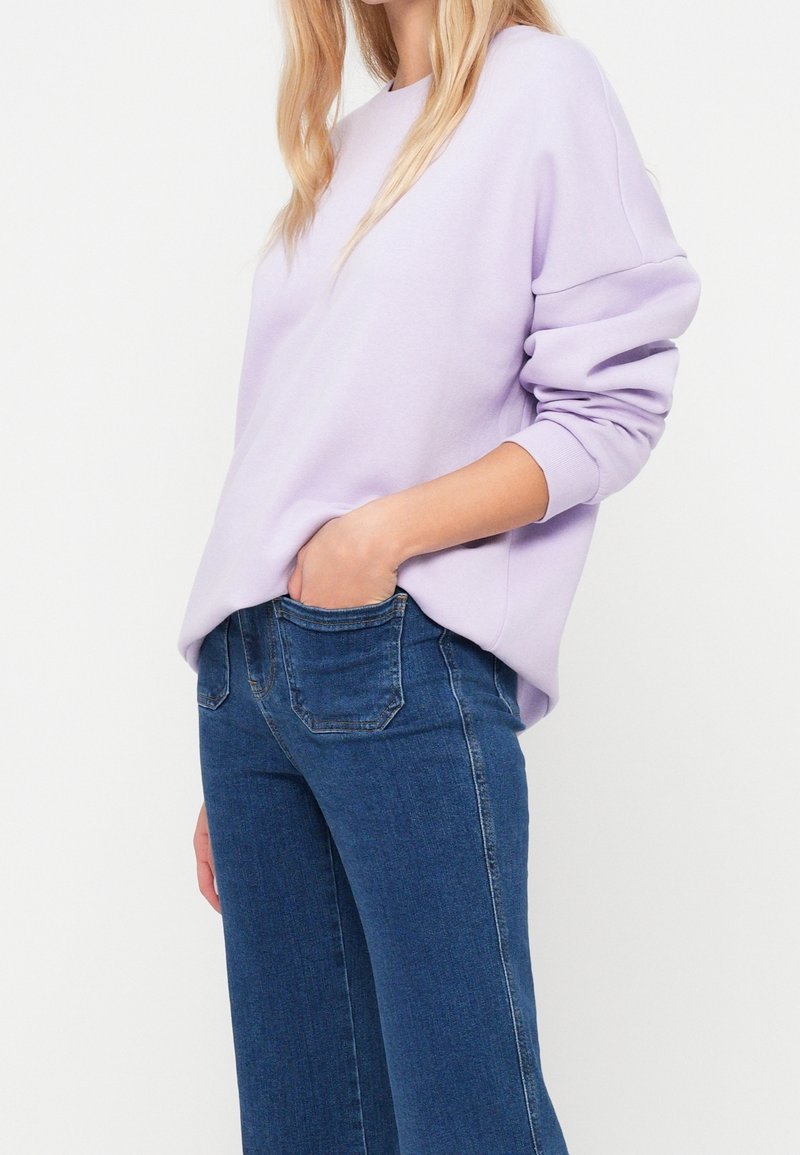 Personne portant un sweat-shirt à manches longues violet clair avec les mains dans les poches d'un jean bleu taille haute sur fond blanc.