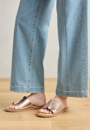 Metalne roza zlate natikače s širokim odprtjem za prste, peščena podlaga in gladka tekstura, v kombinaciji s svetle modre široke hlače.