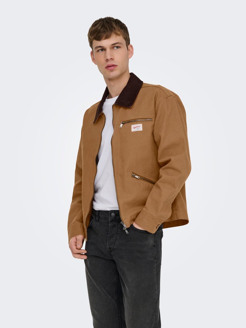 Jeune homme portant une veste marron avec un col noir, une chemise blanche et un jean noir, debout avec une main dans la poche devant un fond uni.