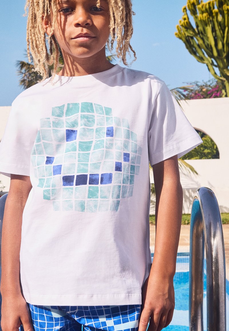 Camiseta blanca con un patrón de mosaico azul y turquesa. El modelo lleva puesta un bañador de cuadros azules a juego. Escalera de metal de piscina al fondo.