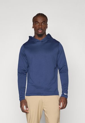 Marineblå langærmet hættejakke med en tekstureret overflade og justerbar hætte. Har et hvidt Nike swoosh på håndleddet. Bæres med beige bukser.