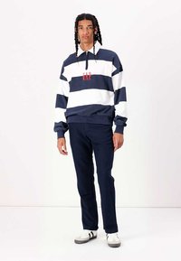 Jersey de rayas azul marino y blanco con cuello con cremallera y logo rojo de "GAP", combinado con pantalones azul marino oscuros y zapatillas blancas con rayas negras.