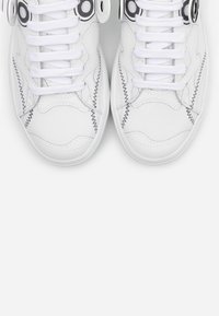 Białe tenisówki z teksturowanym materiałem, z okrągłymi czarnymi akcentami i kontrastującymi szwami. Sznurowadła są białe, a design ma charakter sportowy.