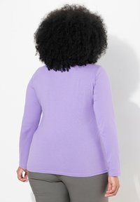 Fialová košile s dlouhými rukávy a přiléhavým střihem, vyrobená z měkkého materiálu. Má zaoblený výstřih a hladký lem, v kombinaci se šedými kalhotami.