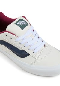 Sneaker din piele albă cu accente în nuanțe de vișină și dunga albastră pe lateral, având șireturi albe și un patch cu logo verde pe limbă.