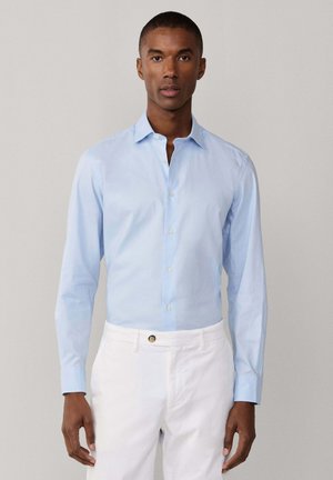 Homme portant une chemise bleu clair à manches longues, boutonnée et rentrée dans un pantalon blanc, debout devant un fond clair uni.