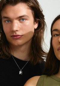Collar de pendant de plata en una modelo con cabello largo y castaño; presenta una forma rectangular y un grabado sutil. La modelo lleva una camiseta negra.