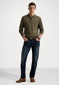 Πουλόβερ πόλο σε ελιά πράσινο, σκούρο τζιν, καφέ δερμάτινα παπούτσια και ρολόι. Η εμφάνιση έχει casual στυλ.