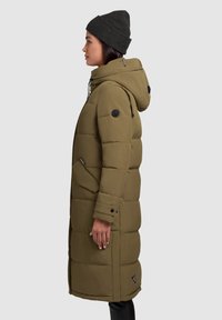 Olive green padded long coat with a hood, zip closure, side pockets, and textured fabric. Features a logo patch on the sleeve.