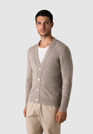 Mężczyzna ubrany w beżowy, prążkowany kardigan z białymi guzikami, na białą koszulę i jasnobeżowe spodnie, stoi na tle gładkiego, jasnego tła.