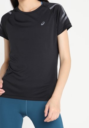 Personne portant un t-shirt de sport Asics à manches courtes noir avec des accents gris et des leggings bleus, debout devant un fond blanc.
