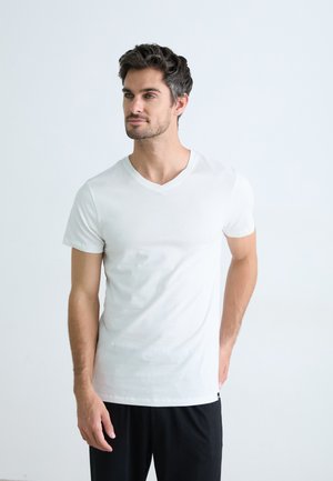 T-shirt en coton blanc à col en V, à manches courtes, texture lisse et coupe ajustée, porté avec un pantalon noir sur un fond clair.