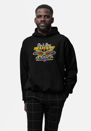 Man wearing black hoodie with colorful "Don't Stop Believin' Journey" text and graphic, paired with black pants with white grid lines.