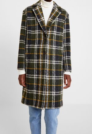 Personne portant un long manteau à carreaux vert foncé avec des rayures blanches, noires et moutarde, un col roulé blanc et un jean bleu clair, debout contre un mur blanc.