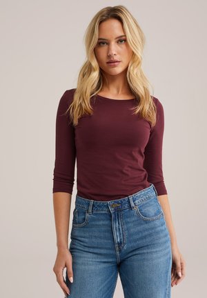Langärmliges Oberteil aus burgunderfarbenem Baumwollstoff mit Rundhalsausschnitt. Kombiniert mit hochtaillierten blauen Jeans aus Denim mit einem ausgewaschenen Finish.