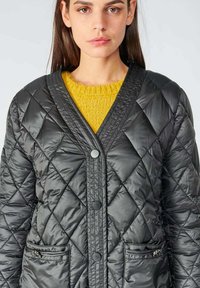 Doudoune noire matelassée avec un col en V, dotée de deux poches avant et de boutons noirs. Elle est portée par-dessus un pull en laine tricoté jaune vif.