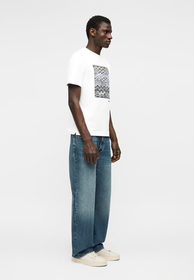 Άνδρας που φοράει λευκό μπλουζάκι με γραφικό σχέδιο, φαρδιά μπλε τζιν και λευκά αθλητικά παπούτσια, στέκεται προφίλ μπροστά σε ένα απλό λευκό φόντο.